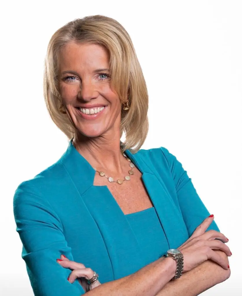 Smiling blonde woman in a teal blazer with arms crossed, wearing a necklace and watch.