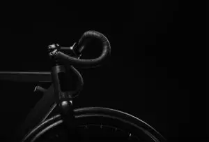Close-up of a black road bike's drop handlebars and front wheel against a dark background, highlighting the sleek frame and components.