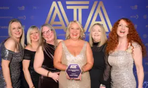 Six smiling women in evening gowns pose together at a blue ATA backdrop, with the woman in the center wearing a gold sequin dress and holding a purple award plaque.