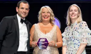 Three people in formal attire pose on stage, with a woman in a glittering gown holding an ATA award plaque.