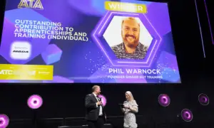 Stage at an awards ceremony with a large screen showing 'WINNER' and 'PHIL WARNOCK FOUNDER GINGER NUT TRAINING' as two presenters speak on the dark stage with purple lighting and circular wall lights behind them.