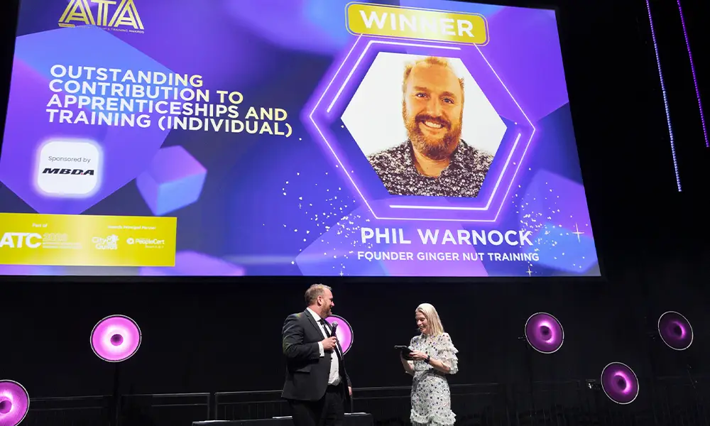 Stage at an awards ceremony with a large screen showing 'WINNER' and 'PHIL WARNOCK FOUNDER GINGER NUT TRAINING' as two presenters speak on the dark stage with purple lighting and circular wall lights behind them.