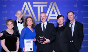 Six people in formal attire pose at an ATA awards backdrop; a woman in blue holds a certificate, a man in the center holds a hexagonal trophy.
