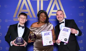 Three formally dressed individuals pose with award certificates against a blue ATA backdrop; middle person holds two plaques while the others hold certificates and trophies, smiling for the camera.