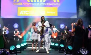 Saxophonist in a white suit performs on stage at the Apprenticeship & Training Awards, with a live band and photographers in the audience.