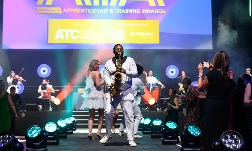 Saxophonist in a white suit performs on stage at the Apprenticeship & Training Awards, with a live band and photographers in the audience.