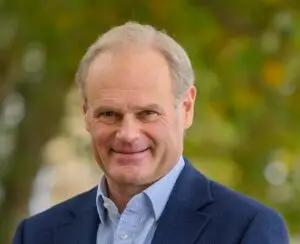 rob - head only headshot | Apprenticeship and Training Awards | Apprenticeship and Training Awards Smiling middle-aged man in a navy blazer and light blue shirt, outdoors with blurred greenery background.