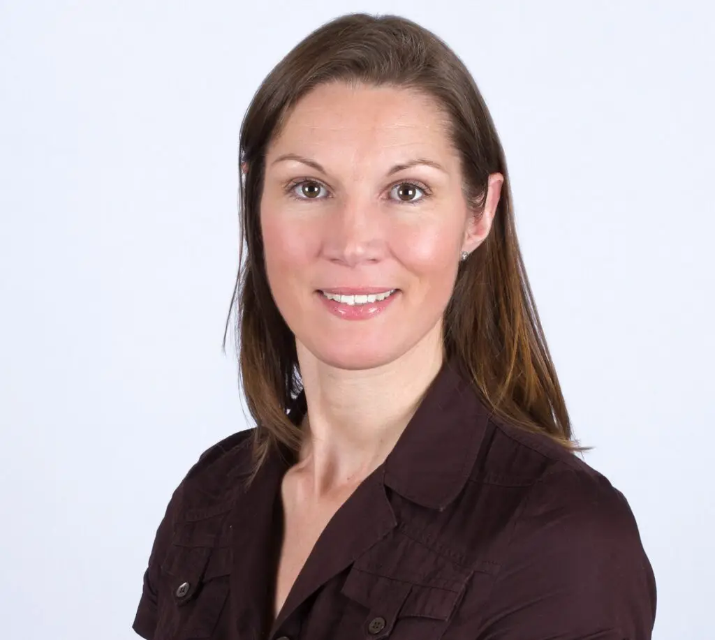 Head-and-shoulders portrait of a smiling woman with brown hair wearing a dark brown shirt against a pale gray background.