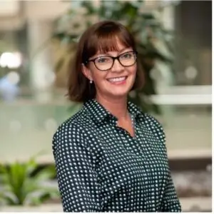 Smiling woman with short brown hair and glasses in a bright office, wearing a dark green polka-dot blouse.
