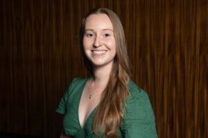 Portrait of a smiling woman with long light brown hair wearing a green blouse, standing in front of a dark wooden backdrop.
